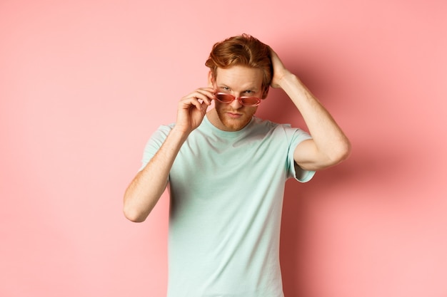 Beau jeune homme rousse à lunettes de soleil, se brosser les cheveux avec la main et à la recherche de suffisance et confiant à la caméra, debout sur fond rose.
