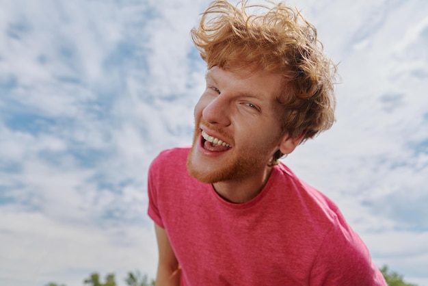 Beau jeune homme rousse regardant la caméra et souriant avec le ciel en arrière-plan