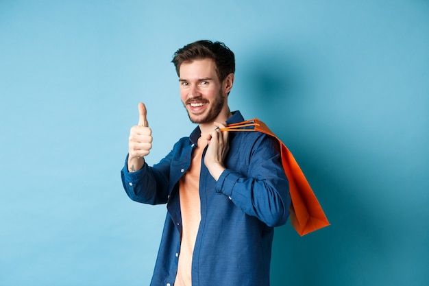 Beau jeune homme avec sac à provisions montrant les pouces vers le haut et souriant, louant une bonne offre de promotion, debout sur fond bleu.