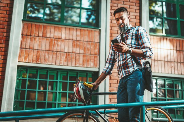 Un beau jeune homme se rend en ville avec son vélo, marche à côté et lit des messages sur un smartphone.