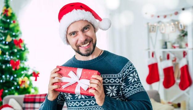 Beau jeune homme souriant barbu dans un bonnet de Noel avec un cadeau de Noël