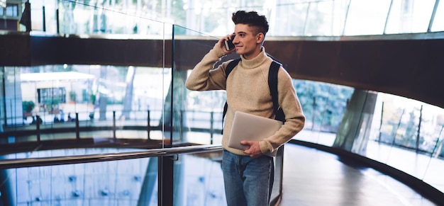 Beau jeune homme souriant dans des vêtements décontractés avec sac à dos parlant au téléphone et portant un ordinateur portable