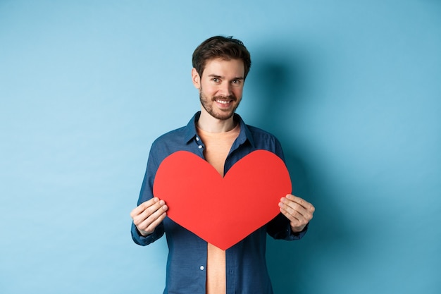 Beau jeune homme souriant, montrant une carte postale de grand coeur rouge pour la Saint-Valentin, regardant la caméra heureuse, debout sur fond bleu.