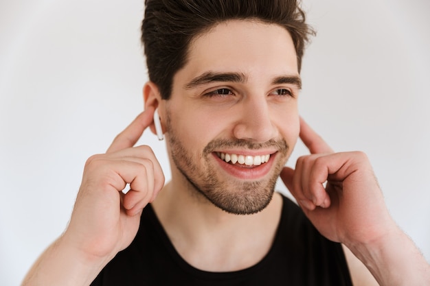 Beau jeune homme sportif souriant et joyeux isolé sur un mur blanc, écoutant de la musique avec des écouteurs.