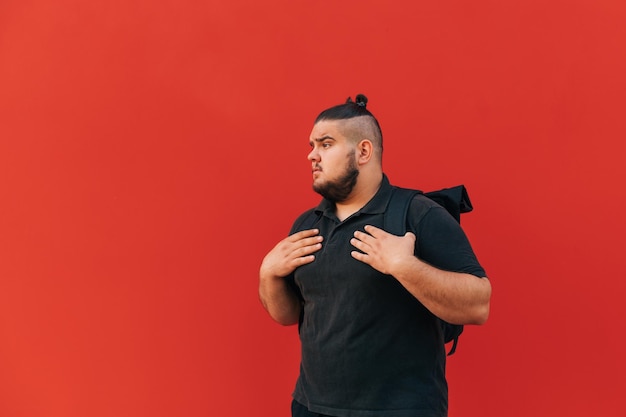 Beau jeune homme en surpoids se tient avec un sac à dos sur le dos sur un fond de mur rouge