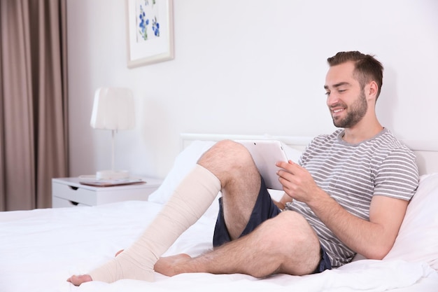 Beau jeune homme avec tablette et jambe bandée assis sur le lit à la maison