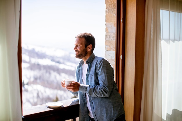 Beau jeune homme avec une tasse de thé chaud à la fenêtre d'hiver