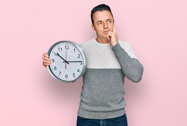 Beau jeune homme tenant une grande horloge visage sérieux pensant à une question avec la main sur le menton réfléchi à une idée confuse