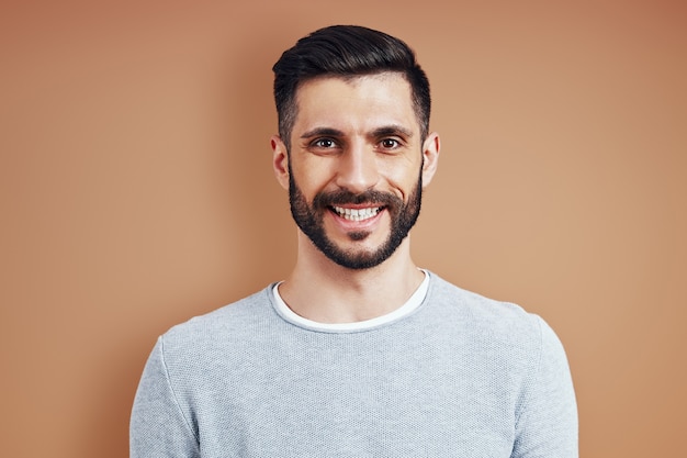 Beau jeune homme en tenue décontractée souriant et regardant la caméra en se tenant debout contre le mur marron