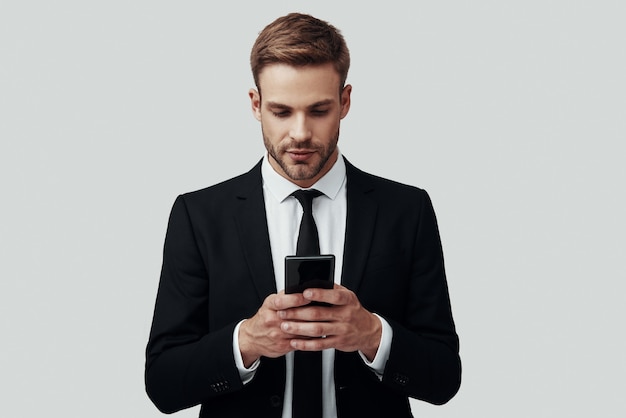 Beau jeune homme en tenue de soirée à l'aide d'un téléphone intelligent en se tenant debout sur fond gris