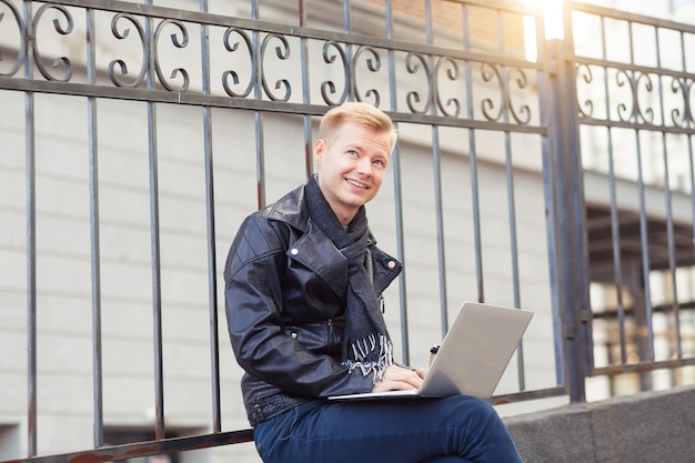 Beau jeune homme travaillant sur ordinateur portable et souriant dans la rue de la ville