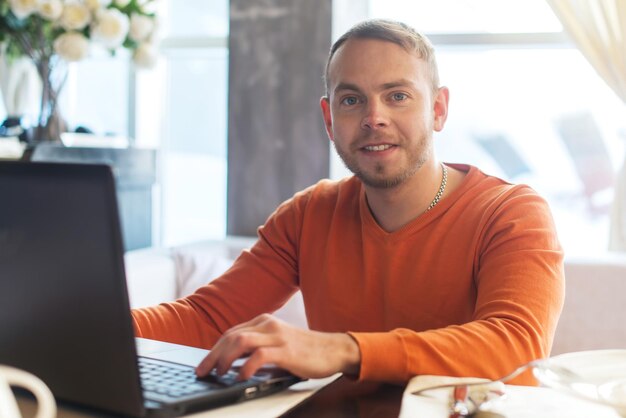 Beau jeune homme travaillant sur ordinateur portable souriant regardant la caméra tout en dégustant un café au café