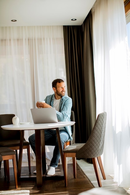 Beau jeune homme utilisant un ordinateur portable et buvant du thé dans le salon