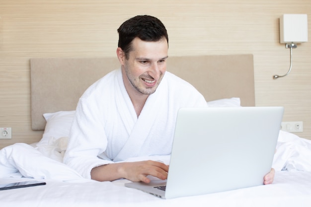 Beau jeune homme utilisant un ordinateur portable en position couchée sur le lit.