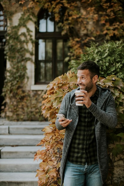 Beau jeune homme utilisant un téléphone portable et tenant un café à emporter dans le parc