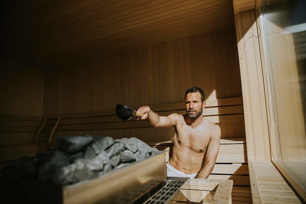 Beau jeune homme versant de l'eau sur des pierres chaudes dans le sauna