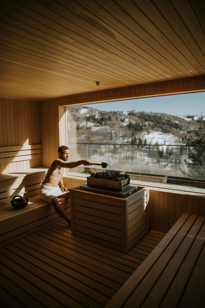 Beau jeune homme versant de l'eau sur des pierres chaudes dans le sauna