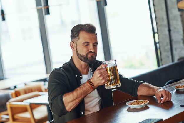 Beau jeune homme en vêtements décontractés appréciant la bière tout en passant du temps au pub