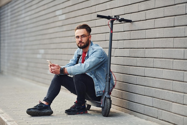 Beau jeune homme en vêtements décontractés assis sur un scooter électrique à l'extérieur pendant la journée ensoleillée