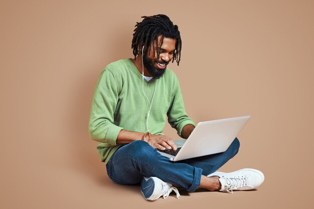 Photo beau jeune homme en vêtements décontractés intelligents utilisant un ordinateur portable et souriant assis contre un mur marron