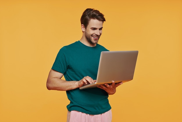 Beau jeune homme en vêtements décontractés utilisant un ordinateur portable et souriant en se tenant debout contre le mur jaune