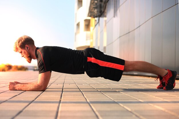 Beau jeune homme en vêtements de sport gardant la position de la planche tout en faisant de l'exercice à l'extérieur