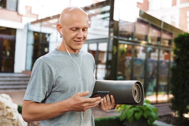 beau jeune homme de yoga positif tenant un tapis de fitness à l'extérieur à l'aide d'un téléphone portable.