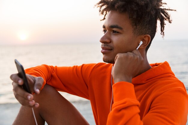 Beau jeune mec émotionnel à l'extérieur sur la plage à l'aide de téléphone portable, écouter de la musique