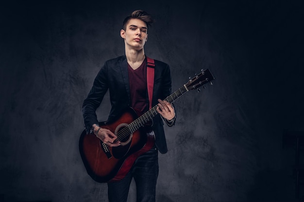Beau jeune musicien aux cheveux élégants dans des vêtements élégants avec une guitare dans ses mains jouant et posant sur un fond sombre.