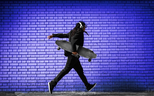 Beau jeune patineur de dreadlocks avec des écouteurs en cours d'exécution dans un costume près du mur bleu dans la rue.