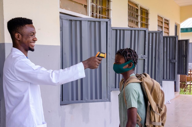 Un beau jeune professeur de classe africain tenant comme thermomètre pour scanner la température de son élève avant d'entrer dans la salle de classe