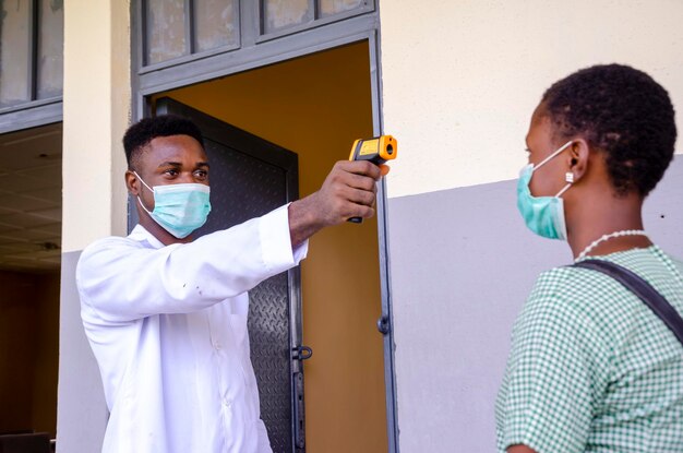 Un beau jeune professeur de classe africain tenant comme thermomètre pour scanner la température de son élève avant d'entrer dans la salle de classe