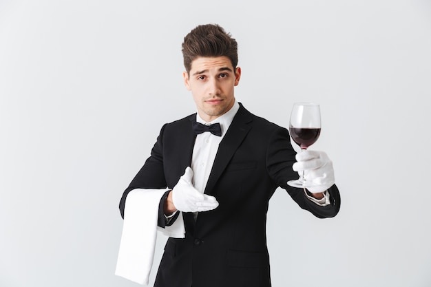 Beau jeune serveur en smoking tenant un verre de vin rouge isolé sur mur gris