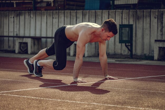 Beau jeune sportif concentré se préparant à commencer à courir sur une piste en tartan sur le stade.