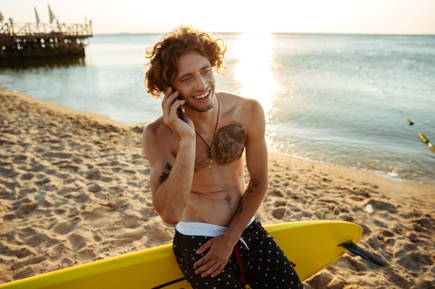 Beau jeune surfeur frisé parlant sur smartphone alors qu'il était assis sur la planche de surf à la plage