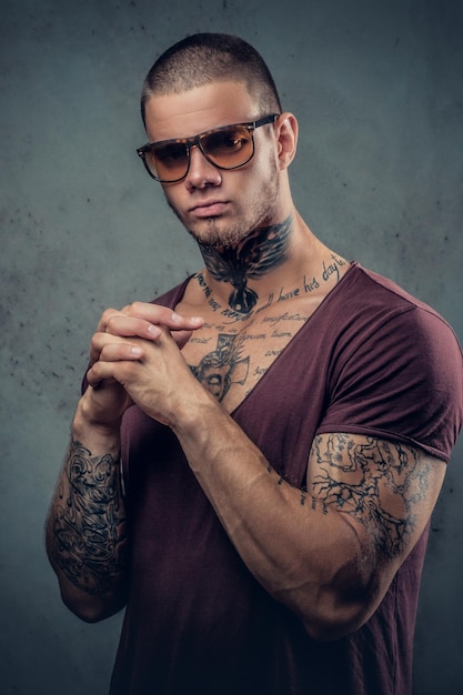 Beau mâle athlétique en lunettes de soleil avec des tatouages sur les bras et le cou posant sur un fond artistique gris dans un studio.