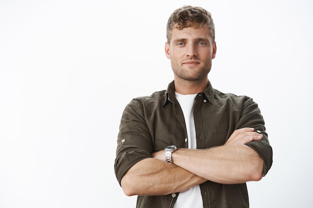 Beau mâle blond masculin confiant avec des poils et des yeux bleus, tenant les mains croisées sur la poitrine dans une pose sûre d'elle, souriant narquoisement ravi, étant assuré de son propre succès.