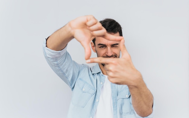 Beau mâle heureux barbu cligner des yeux et regardant à travers un cadre formé par ses mains Homme séduisant faire cadre par doigts isolé sur fond blanc avec espace de copie pour le texte Concept de personnes