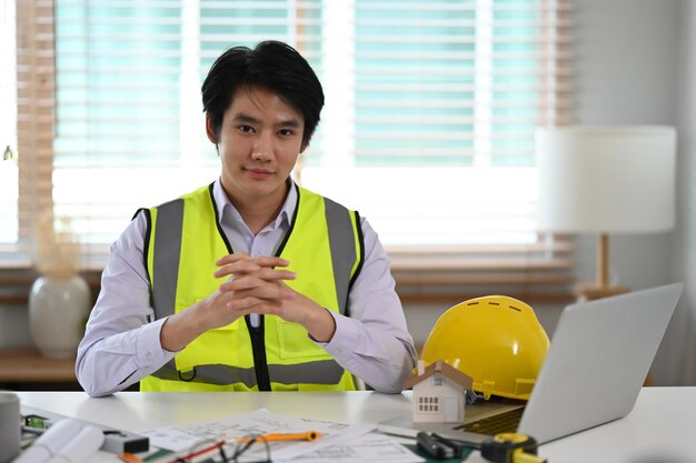 Beau mâle ingénieur en construction en chemise blanche et gilet jaune assis sur le lieu de travail et souriant à la caméra