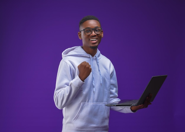 Beau mâle à lunettes en souriant. heureux étudiant africain extatique tenant un ordinateur portable portant un sweat à capuche