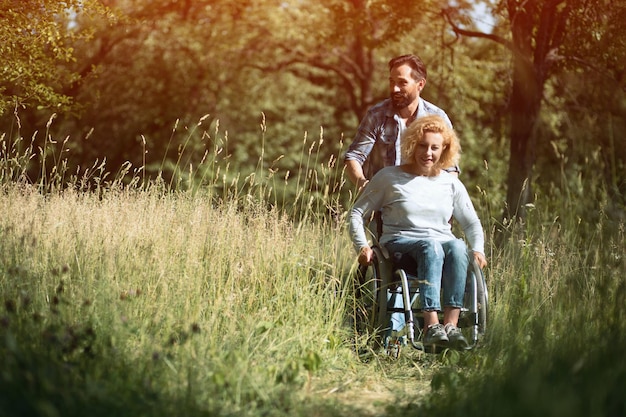 Beau mari déplace le fauteuil roulant avec sa femme souriante dans le parc