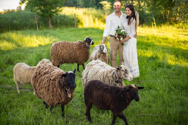 Beau marié élégamment vêtu avec une barbe en lunettes de soleil noires embrasse sa femme La mariée dans une robe avec un bouquet met un chapeau et rit Mariage rustique dans le style de boho au ranch