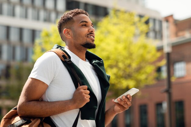 Beau mec afro-américain à l'aide d'un smartphone à l'extérieur marchant dans une zone urbaine après les cours