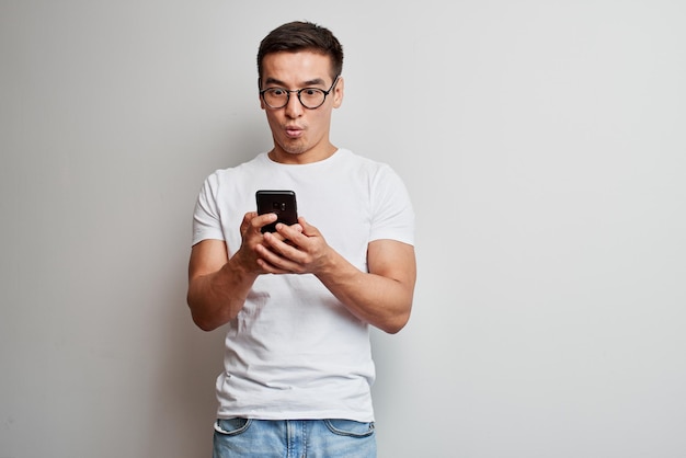 Beau mec asiatique en studio blanc semble surpris par l'écran du téléphone portable. Un homme kazakh choqué dans des lunettes habillées de manière décontractée tient un smartphone dans les mains