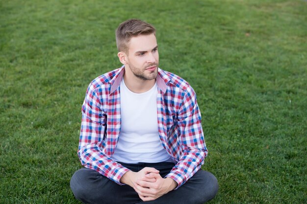 Beau mec assis sur l'herbe verte. Homme mal rasé visage humeur paisible. Penser à l'avenir. Crise existentielle. Étudiant relaxant sur le pré. Étudiant rêveur. Beau mec étudiant à l'extérieur de l'espace de copie.