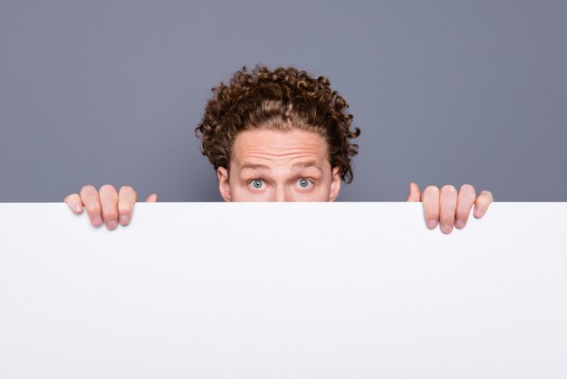 Beau mec attrayant avec des cheveux ondulés se cachant derrière un tableau promotionnel blanc