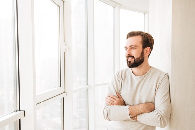 Beau mec ayant barbe et moustache, souriant et regardant de côté par la fenêtre tout en se reposant avec les mains jointes à la maison