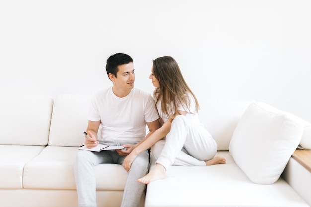 Beau mec et belle fille amoureuse se regardent dans les yeux, assis sur un canapé blanc et doux dans une pièce minimaliste moderne et lumineuse. Le jeune homme prend des notes avec un stylo dans son cahier.