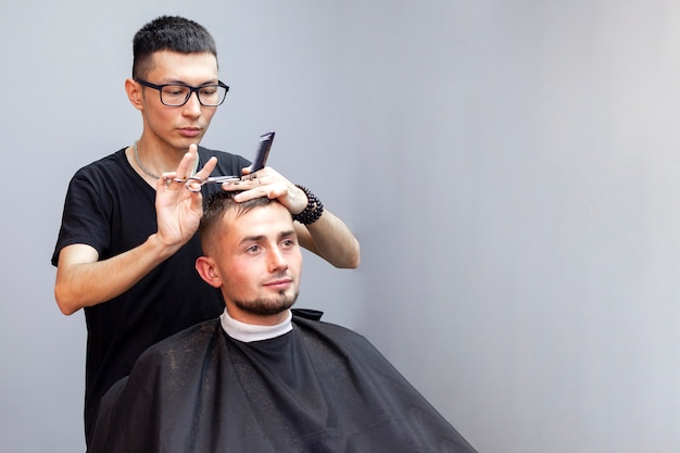 Beau mec a une coupe de cheveux chez le coiffeur, un jeune coiffeur kazakh coupe manuellement avec des ciseaux et un peigne, le maître fait une coupe courte contre un mur gris