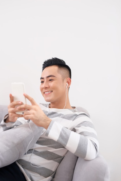 Beau mec dans les écouteurs utilise un téléphone intelligent et sourit en position couchée sur le canapé à la maison